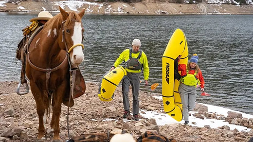 Two people wearing drysuits carrying kayaks from a lake in winter